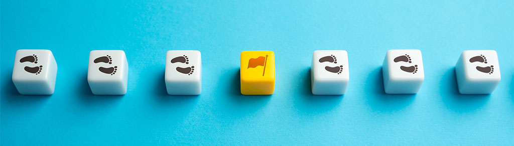 A row of seven dice on a vibrant blue background. All are white with footprints in black, except for the fourth die, which is yellow and features a flag. This image symbolizes the achievement of a goal. This visual metaphorically suggests setting and accomplishing small, incremental goals in a process or journey.