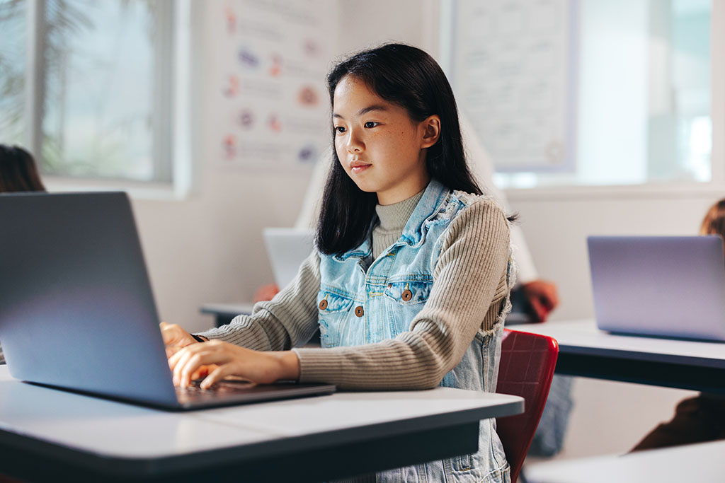 A young Asian student is intently working on a laptop in a classroom setting. She is wearing a denim jumper over a long-sleeved shirt and is seated at a white desk. The background is softly focused, with educational posters visible on the wall and other students also working on laptops. The image conveys a sense of focus and concentration, typical of an academic or learning environment where technology and computers are integral tools.