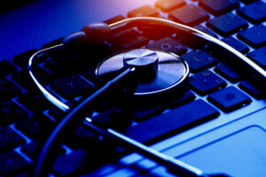 A close-up of a stethoscope resting on a laptop keyboard, illuminated by blue light. The stethoscope's diaphragm is centered and in focus, implying a metaphorical diagnosis or maintenance of computer health