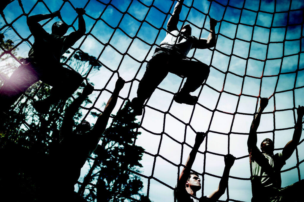 Silhouetted figures against a bright sky, climbing on a giant rope net in an outdoor setting. The ropes crisscross to create large squares. The climbers are at various points on the net, grasping and stepping on the ropes, suggesting a military exercise.